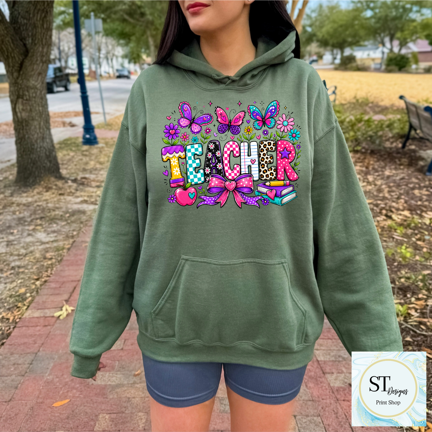 Colorful teacher graphic crewneck sweatshirt from ST Designs Store, featuring vibrant school-themed illustrations on a sand-colored fabric.